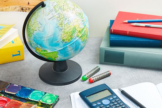 Schreibtisch mit Globus, Taschenrechner, Tuschkasten, Englischwörterbuch, Bücher, Heften und Stiften.