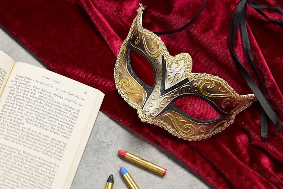 Tisch auf dem ein Textbuch, Schminkstifte, eine Goldene Maske und roter Samtvorhang