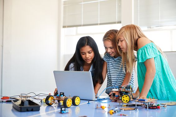 Drei Mädchen stehen vor einem Notebook-Computer. Auf dem Tisch liegen viele Teile von Roboterbausätzen.