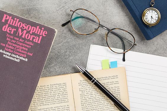 Auf dem Schreibtisch liegen Philosophiebücher, eine Brille, eine kleine Taschenuhr, Zettel und Stift.