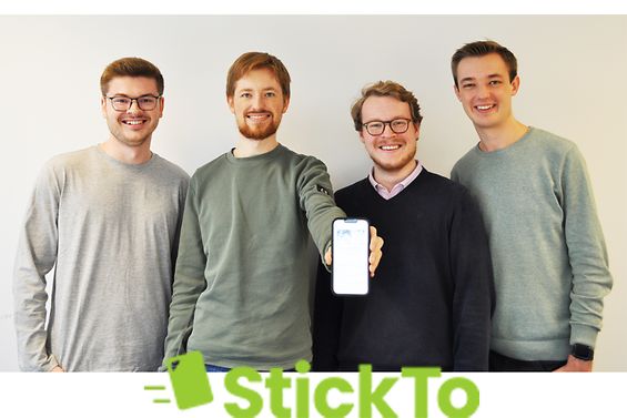 Vier junge Männer stehen vor einer Wand nebeneinander und einer hält ein eingeschaltetes Smartphone in die Mitte.
